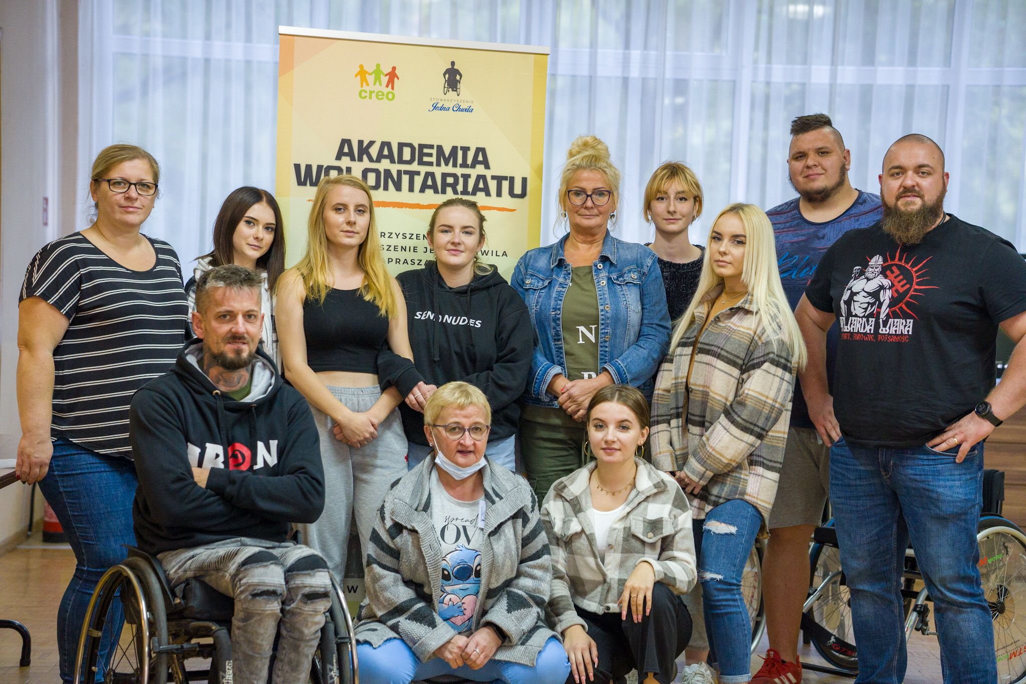 Grupa 12 osób, w tym jedna na wózku, pozuje do zdjęcia. W tle roll up z napisem Akademia Wolontariatu.